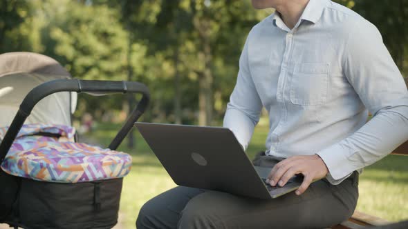 Unrecognizable Young Father Using Laptop and Rocking Baby Stroller in Summer Park Outdoors. Busy