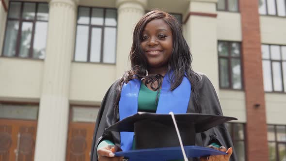 An AfricanAmerican Graduate Woman in a Black Master's Gown Cheerfully Holds a Diploma and a Square