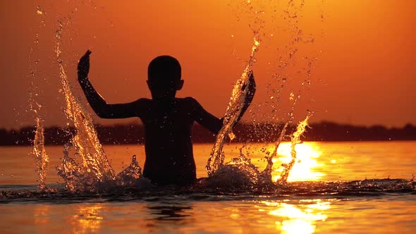 Silhouette of Boy at Sunset Raises Hands and Creating Splashes of Water. Slow Motion