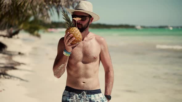 Guy Walking On Tropical Beach With Pineapple. Man Relaxing On Caribbean Beach. Tanned Man In Hat.