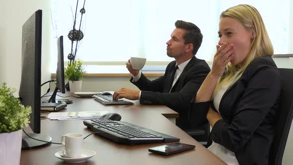 Two office workers, man and woman, work on computers and get tired