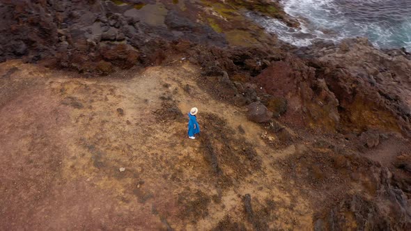 Aerial View of Woman in a Beautiful Blue Dress and Hat Stands on Top of a Mountain in a Conservation