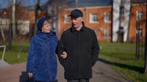 an Elderly Couple Walks Arm in Arm and Talks Against the Background of Blurred Lowrise Houses on a