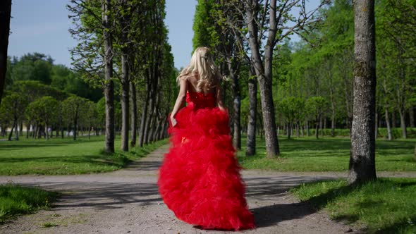 a Blonde in a Red Fluffy Dress Runs Along the Path in the Park