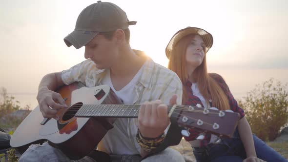 Hipster Young Guy Playing Guitar and His Girlfriend Listening with Interest on the Background