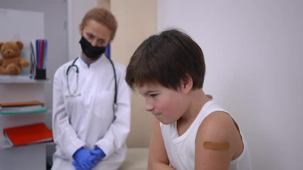 Unsatisfied Caucasian Boy with Medical Patch on Shoulder Looking Back at Blurred Doctor in