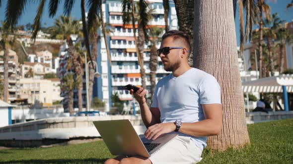 Handsome Man Sitting on the Grass in the City with a Laptop and Talking on the Phone Job Search Rest