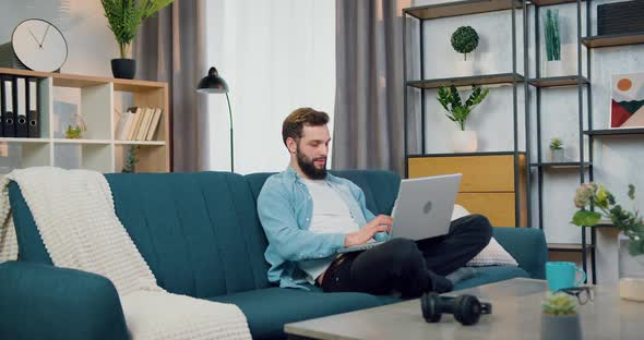 Man Chatting Online with Friends on Laptop while Sitting in Lotus Pose on the Couch at Home