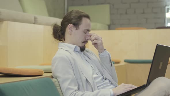 Side View of Tired Caucasian Office Worker Sitting with Laptop and Rubbing Eyes. Exhausted Young Man