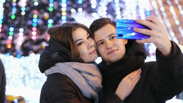 Happy Couple Makes a Selfie on a Modern Phone, Smiling and Kissing in New Year's Night