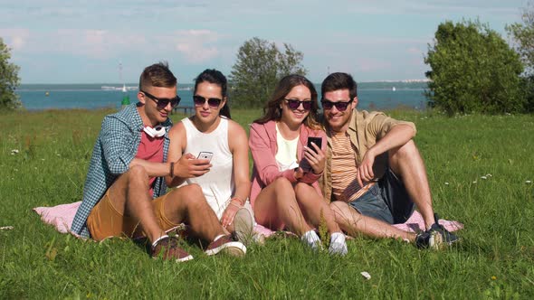 Smiling Friends with Smartphones Sitting on Grass 