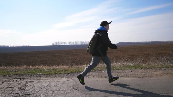 Close Up of Male Child in Cap Jogs Near Plowed Field at Early Spring. Small Kid with Backpack