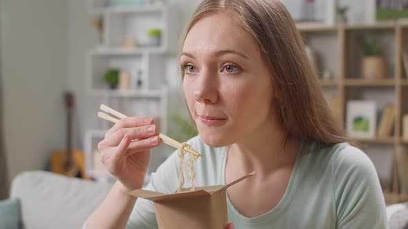 Young Woman Eating Noodles From a Box with Chopsticks at Home on a Couch