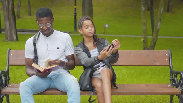 Concentrated Man in Eyeglasses Reading Book As Cheerful Girl Taking Selfie in Sunny Park. Portrait