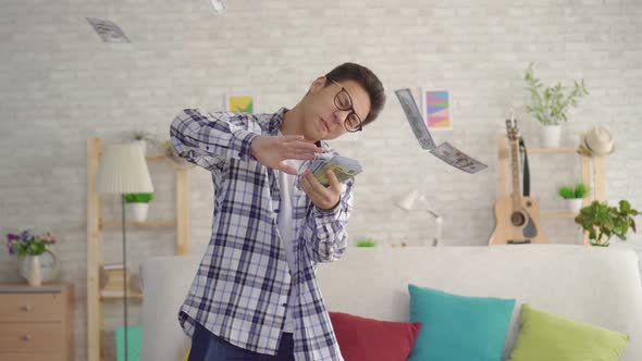 Positive Young Asian Man with Glasses in the Living Room of the House Throws Banknotes