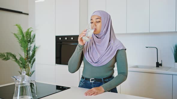 Modern Muslim Young Woman in Jeans and a Hijab Drinks Clean Water While Standing in Her Kitchen