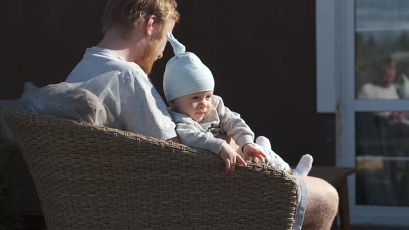 baby sits on dad in a chair on the terrace on a sunny day