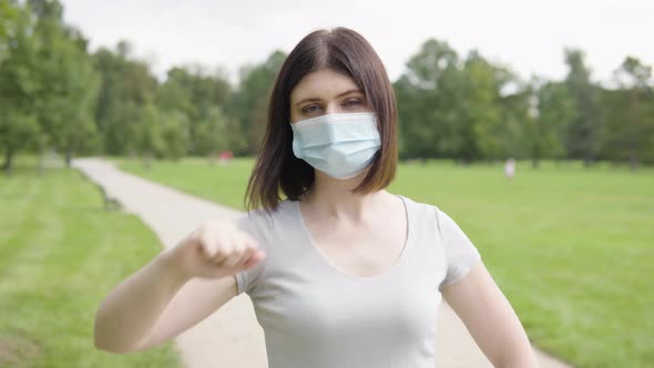 A Young Caucasian Woman in a Face Mask Shows a Thumb Down to the Camera and Shakes Head on a Pathway