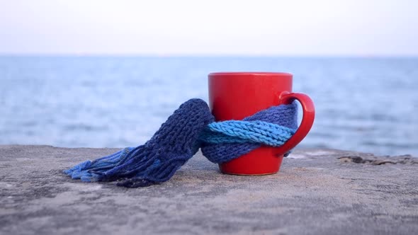 Red Cup with Hot Coffee Tea Tied with Blue Knitted Scarf Stands on Stone