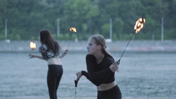 Two Young Women Performing a Show with Flame Balls Standing on the Riverbank