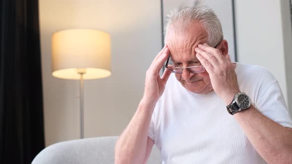 An Old Grayhaired Retired Grandfather is Sitting on the Sofa Massaging His Head and Coughing