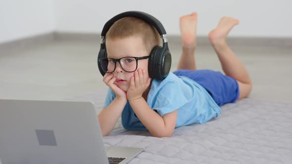 Baby Child Boy in Blue Clothes Lying on Floor in Headphones Watching Laptop