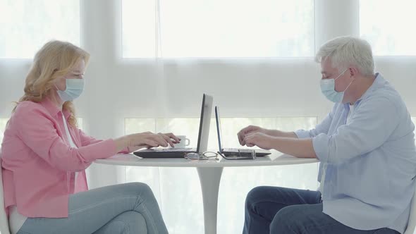 Mid-adult Man and Woman in Face Masks Sitting at the Table and Typing on Laptop Keyboards. Side View