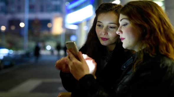 Two young beautiful caucasian women friends outdoor in the city night using smart phone