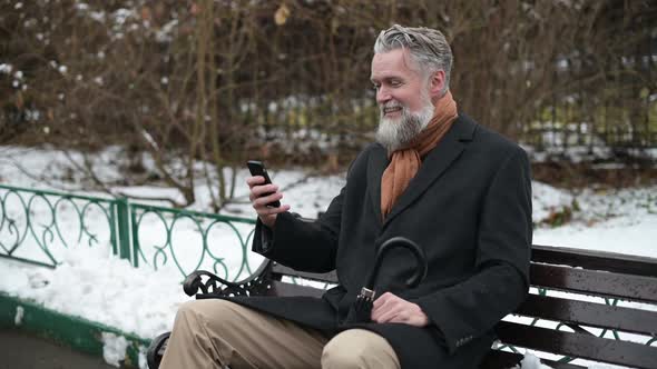 A solid gray-haired man sits on a park bench and talks via video link