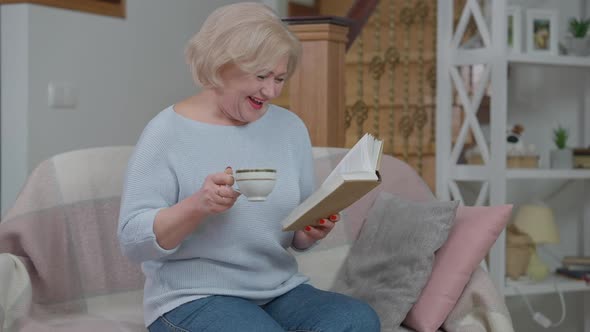 Joyful Senior Beautiful Woman Laughing Reading Book Drinking Tea at Home in Living Room