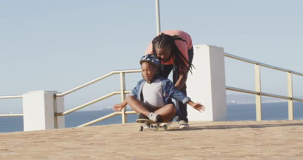 Video of happy african american father learning son how to skateboard on promenade