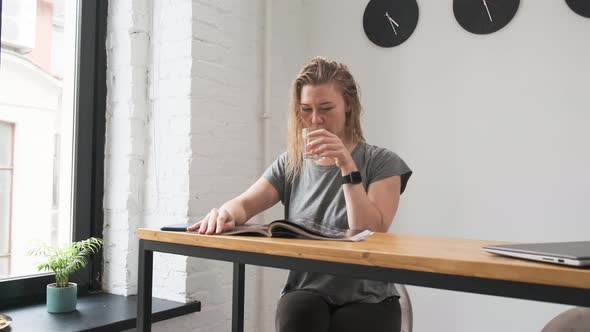 Footage Beautiful Blonde in Gray T-shirt Sits at Table and Reads Magazine. Drinks Water From