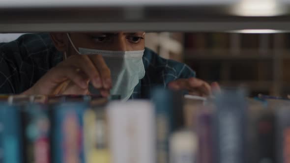 Closeup Hispanic Guy Student in Protective Medical Mask Looking for Right Book on Bookshelf in