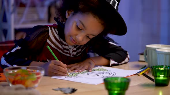 Little African American Girl in Festive Costume and Witch Hat Draws Pumpkin with Pencils