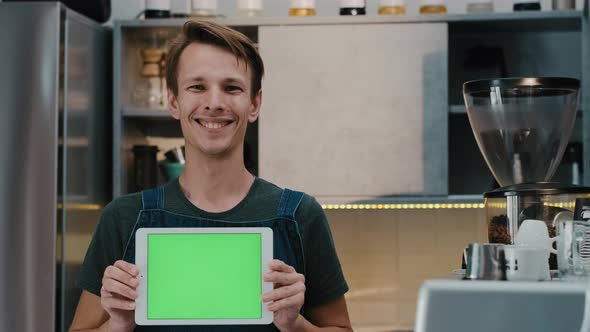 Happy man barista happy  looking at camera showing green screen tablet