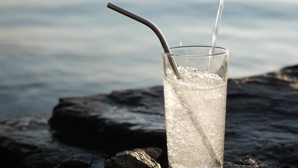 Soda Water Pours Into Tall Glass with Reusable Cocktail Tube on Rocky Seashore