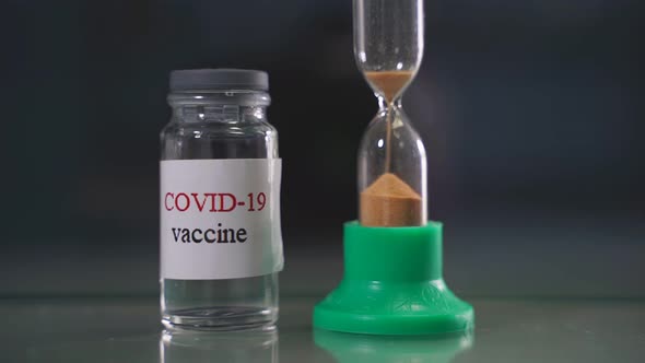 Vials of Coronavirus Vaccine and an Hourglass in the Foreground