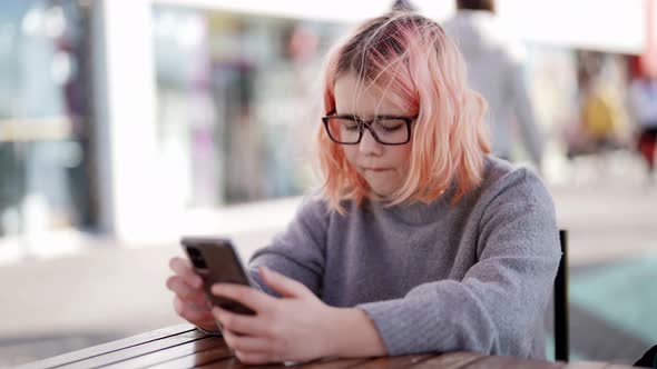 A Teenage Girl with Glasses is Sitting at a Table in a Street Cafe and Playing Video Games on Her