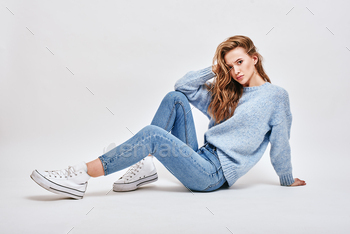 aring blue sweater, jeans, converse, sitting on the floor, touching her hair with a surprised look, isolated over white background
