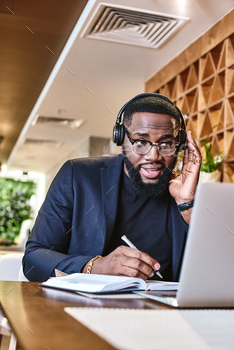 jacket and glasses sitting at the table, indoors, wearing headphones, working with his laptop computer. Young African entrepreneur is listening to his boss, making notes in his notebook. Modern interior