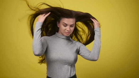 Slender Woman in Gray Turtleneck Stands Against Yellow Background in Studio and Straightens Her Hair