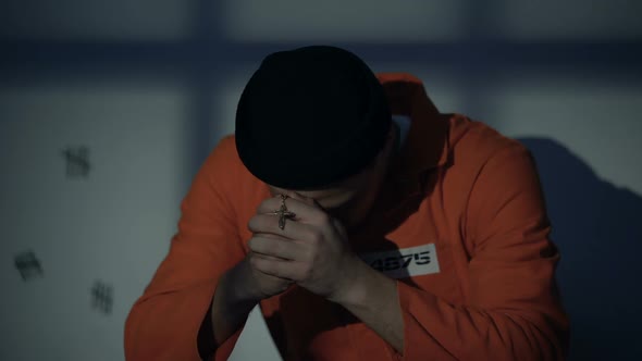 European Imprisoned Male Holding Silver Cross and Praying in Cell Feeling Guilty