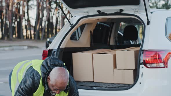 Closeup Mature Delivery Service Worker Unloading Cardboard Boxes From Trunk of Car Elderly African