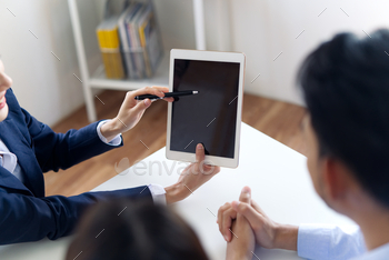  an Asian friendly smiling young couple seated at her desk in the office. The couple looking agent explaining on the tablet carefully .