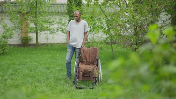 Wide Shot of Thoughtful Adult African American Caregiver Standing at Wheelchair on Summer Backyard