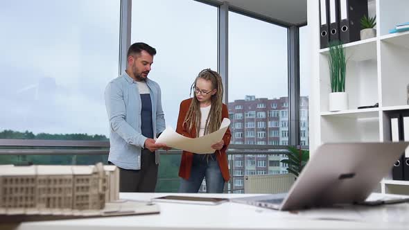 Man and Woman with Dreadlocks Standing Near Design Office Window and Discussing Blueprint