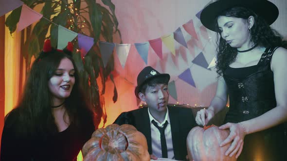 Two Girls and a Guy in Creepy Halloween Costumes are Sitting at a Table Chatting and Carving Faces