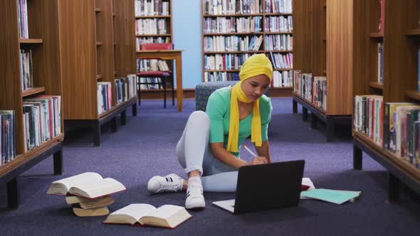 An Asian female student wearing a yellow hijab studying in a library and using laptop