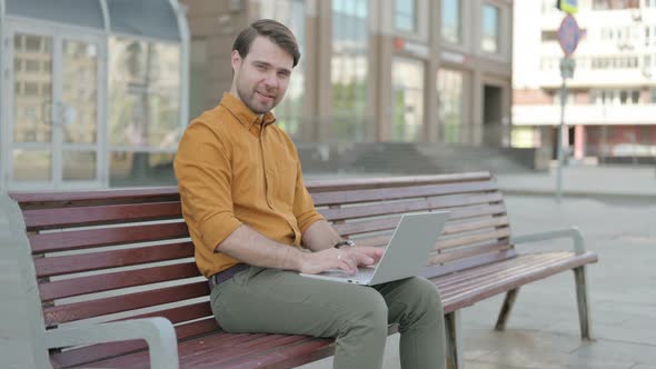 Young Man with Laptop Showing Thumbs Up Sign While Sitting Outdoor on Bench