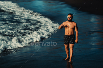 a strong man in black shorts and glasses walks along the beach with black volcanic sand on the